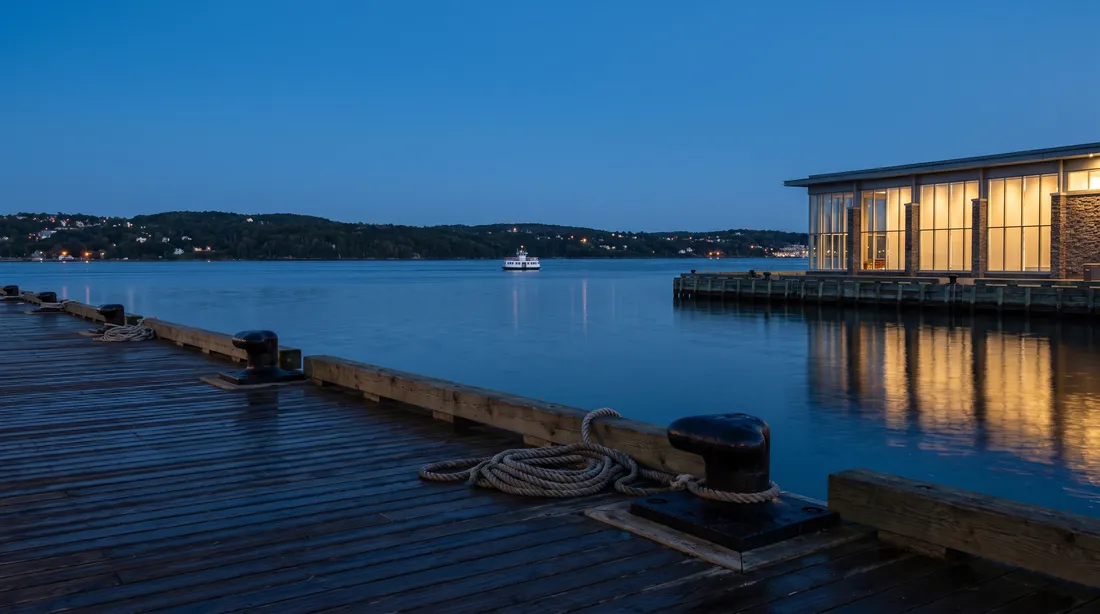 A wooden harbour boardwalk at blue hour with iron bollards and coiled rope, calm water, a lit glass building on the right and a small white ferry far out.